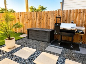 A backyard with a wooden fence, a barbecue grill, a storage chest, and a potted plant.