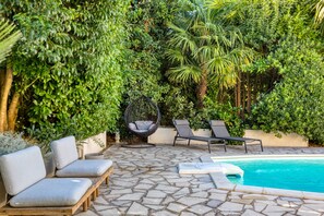 A poolside area with stone flooring, lounge chairs, a hanging chair, and lush greenery. - Holiday Home 'Maison Sur La Mare' with Mountain View, Private Pool and Wi-Fi (Villemagne-l'Argentière)