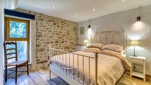 Une chambre avec un mur en pierre, une chaise en bois, un cadre de lit en métal, une table de chevet avec une lampe et une fenêtre.