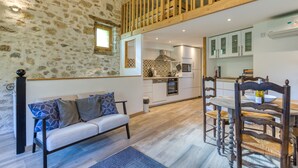 Un intérieur chaleureux avec un mur en pierre, un plafond en bois, un canapé agrémenté de coussins à motifs, une table à manger avec des chaises et un coin cuisine.