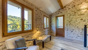 Une pièce aux murs de pierre, au plafond en bois, avec un banc, une chaise et de grandes fenêtres donnant sur un paysage verdoyant.
