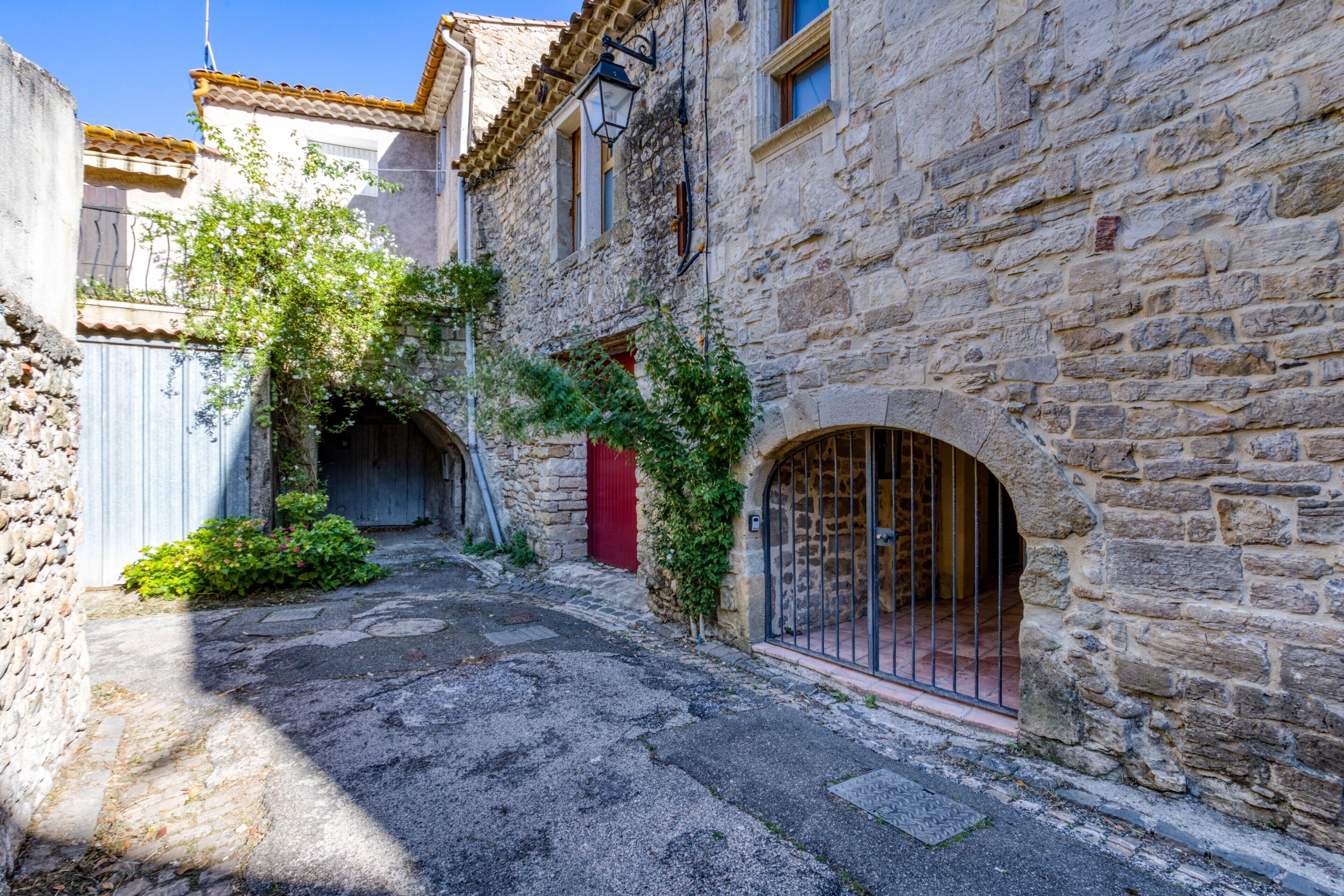 Une ruelle étroite pavée de pierres, avec une porte rouge et un portail en métal, bordée de bâtiments en pierre dotés de fenêtres en arc de cercle.