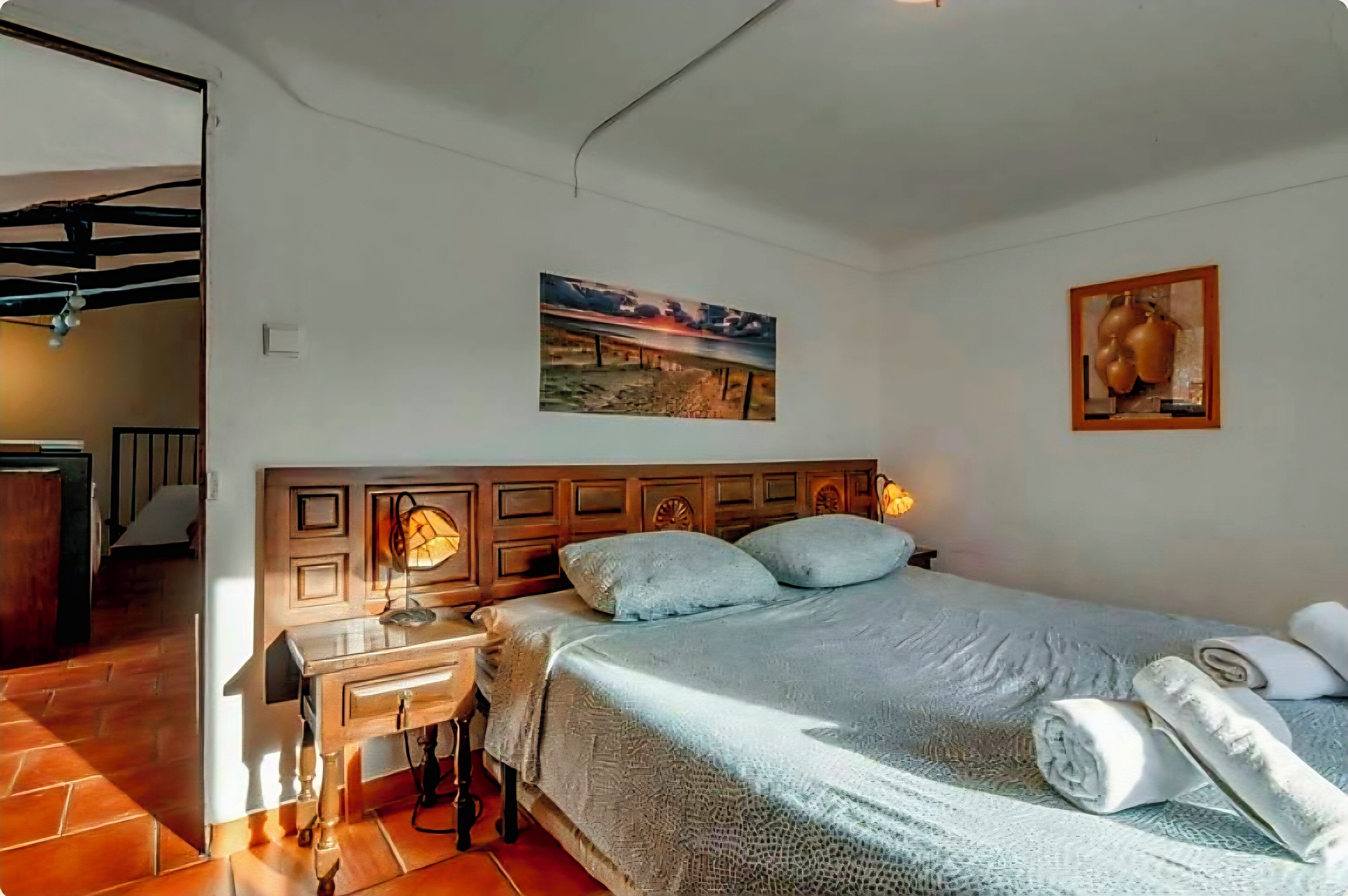 A bedroom with a wooden headboard, a bed with a patterned bedspread, a nightstand with a lamp, and a painting on the wall.