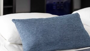 Een bed met een blauw kussen en een gewatteerde deken, een witte muur en een plank met opbergdozen met een patroon en een potplant.