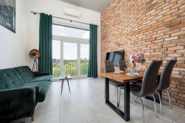 A modern living space with a green sofa, brick wall accents, and a dining table with dark chairs.