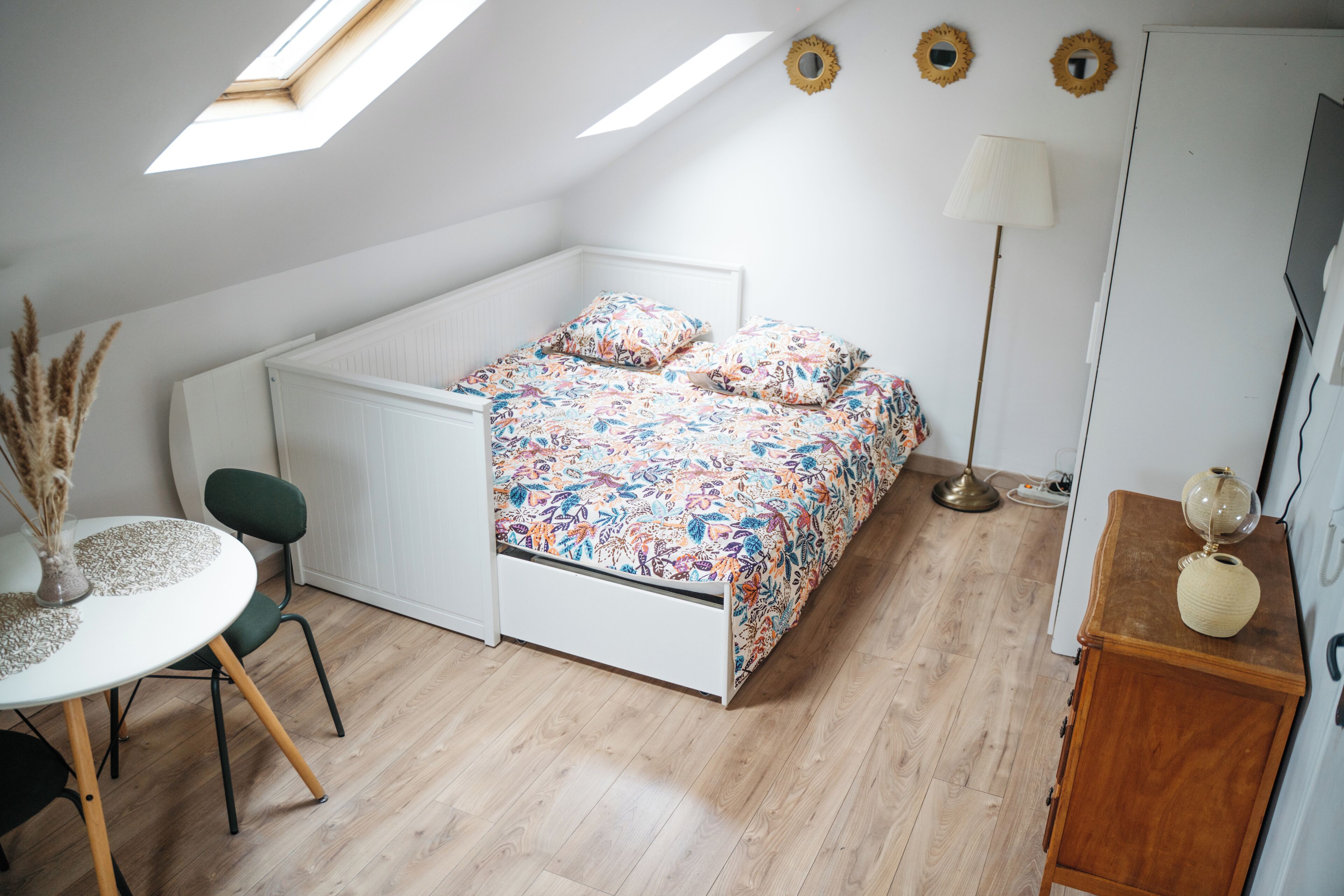 A compact bedroom with a bed, a desk, and a chair. There is a skylight above the bed, a lamp, and a small cabinet.