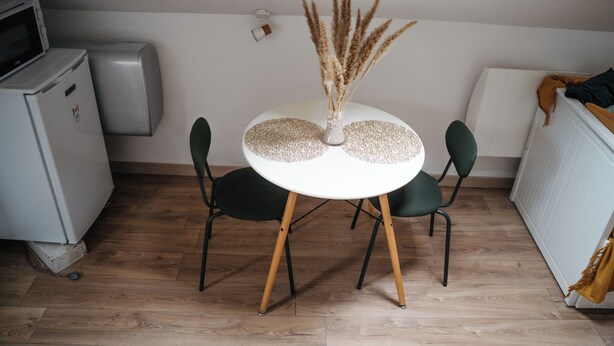 Ein kleiner Essbereich mit einem runden Tisch, zwei Stühlen, einer weißen Vase mit Trockenpflanzen, einer Mikrowelle und einem Heizkörper.