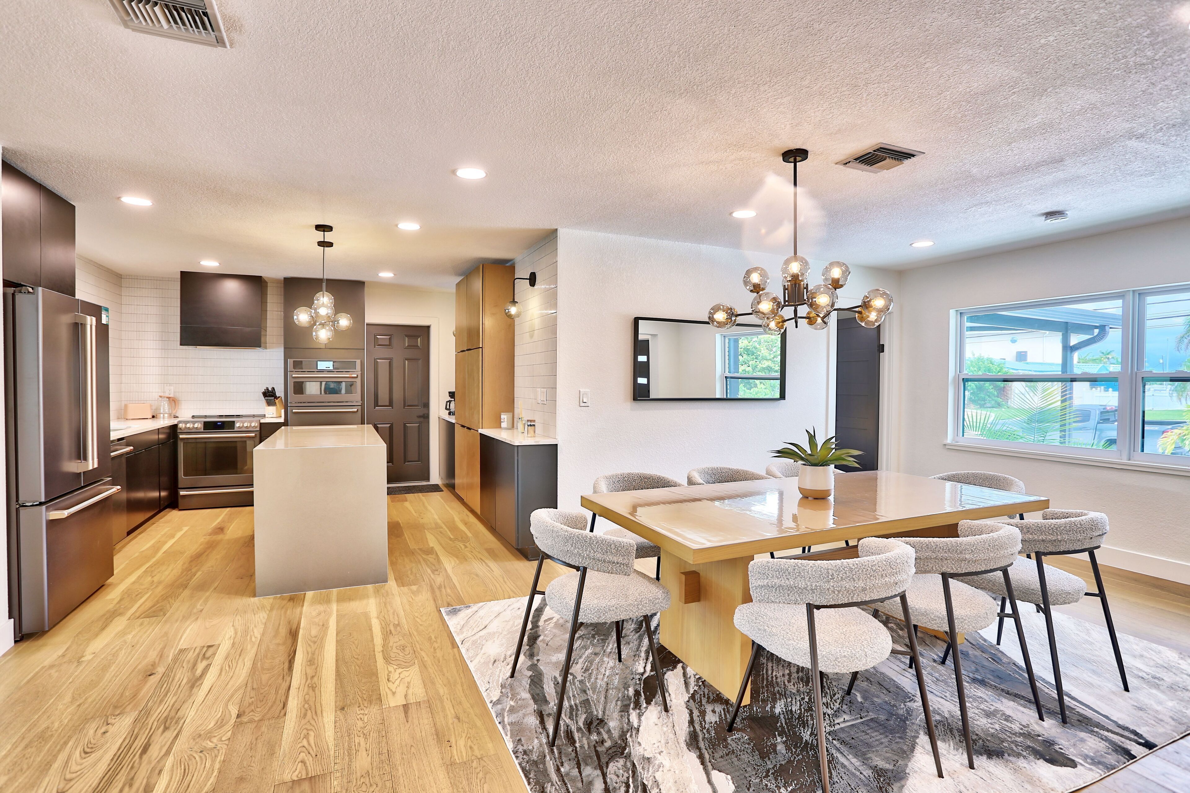 A modern kitchen with a central island, a dining area with a large table and chairs, and a large window providing a view of the outdoors.