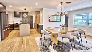 Una cocina moderna con una isla central, un comedor con una gran mesa y sillas, y un amplio ventanal con vistas al exterior.