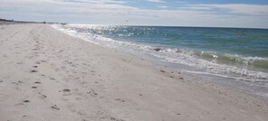 Ein sandiger Strand mit Fußspuren, die zum Wasser führen, ein klarer Himmel und Gebäude in der Ferne.