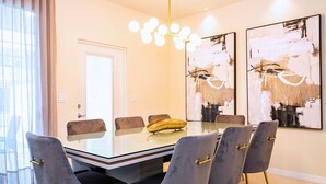 A dining area with a glass table, grey upholstered chairs, and abstract wall art. - Upstay Family Fun 5BR Retreat w Pool & Games Room (Clermont)