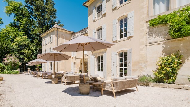 A courtyard with outdoor seating and umbrellas, surrounded by a historic building with large windows and shutters. - Château Saint Perret (Avignon)