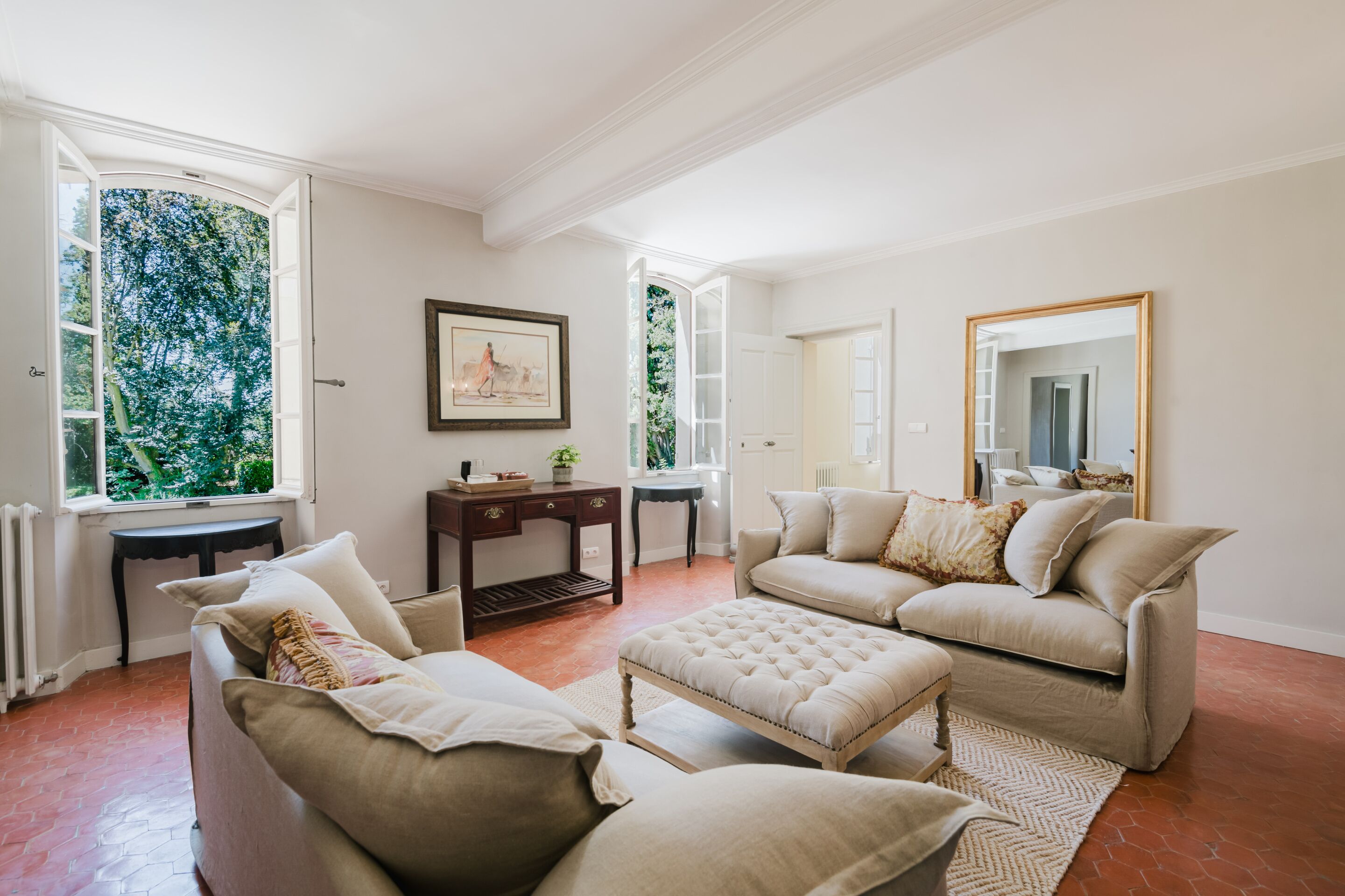 Un salon spacieux avec un canapé beige, un pouf assorti et une table basse. Un tableau encadré orne le mur, ainsi qu’une petite table d’appoint et une fenêtre donnant sur un paysage verdoyant.