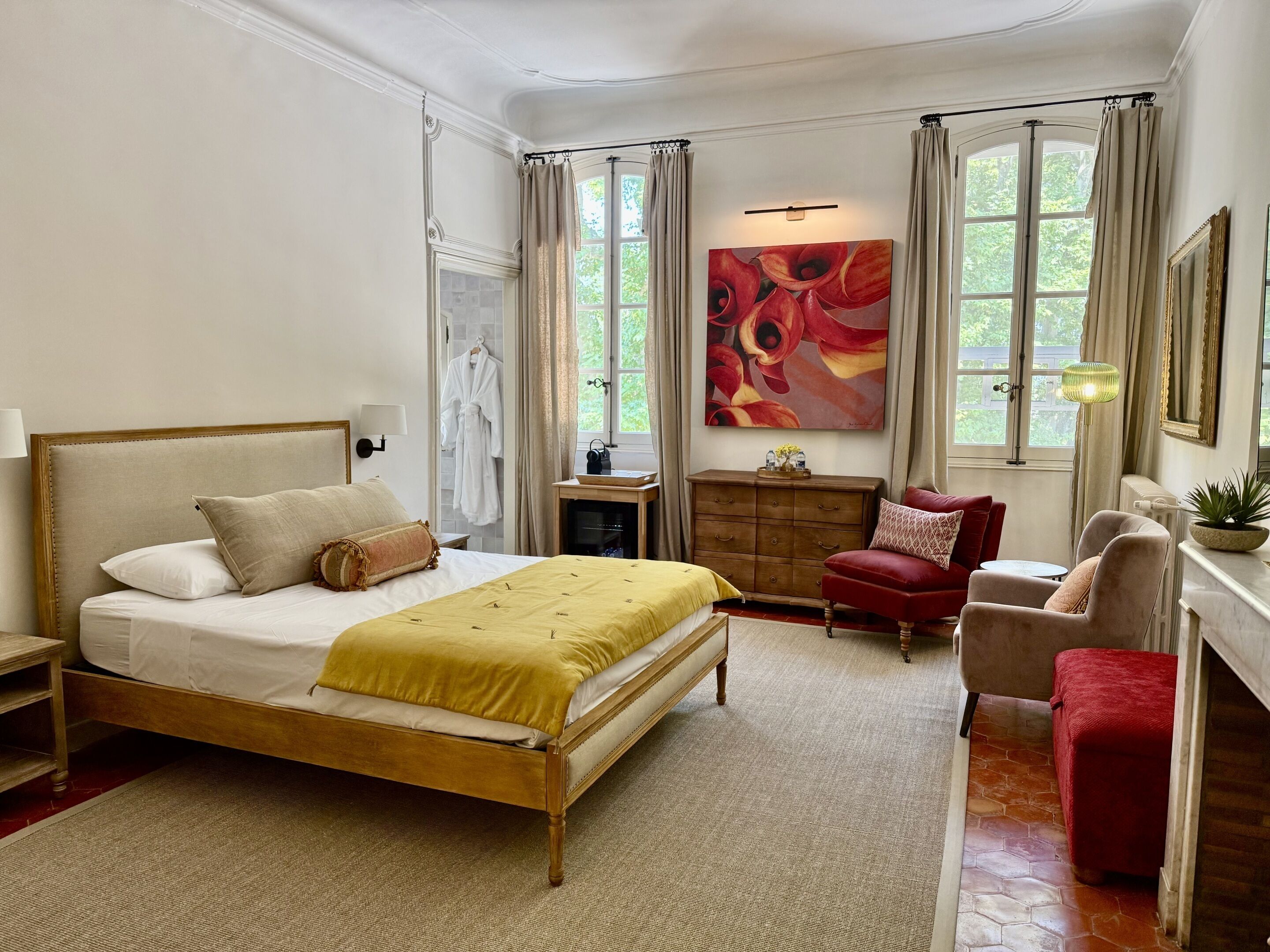 Une chambre bien aménagée, dotée d’un grand lit, d’une commode en bois, d’un coin salon avec des fauteuils et d’une grande fenêtre avec des rideaux.