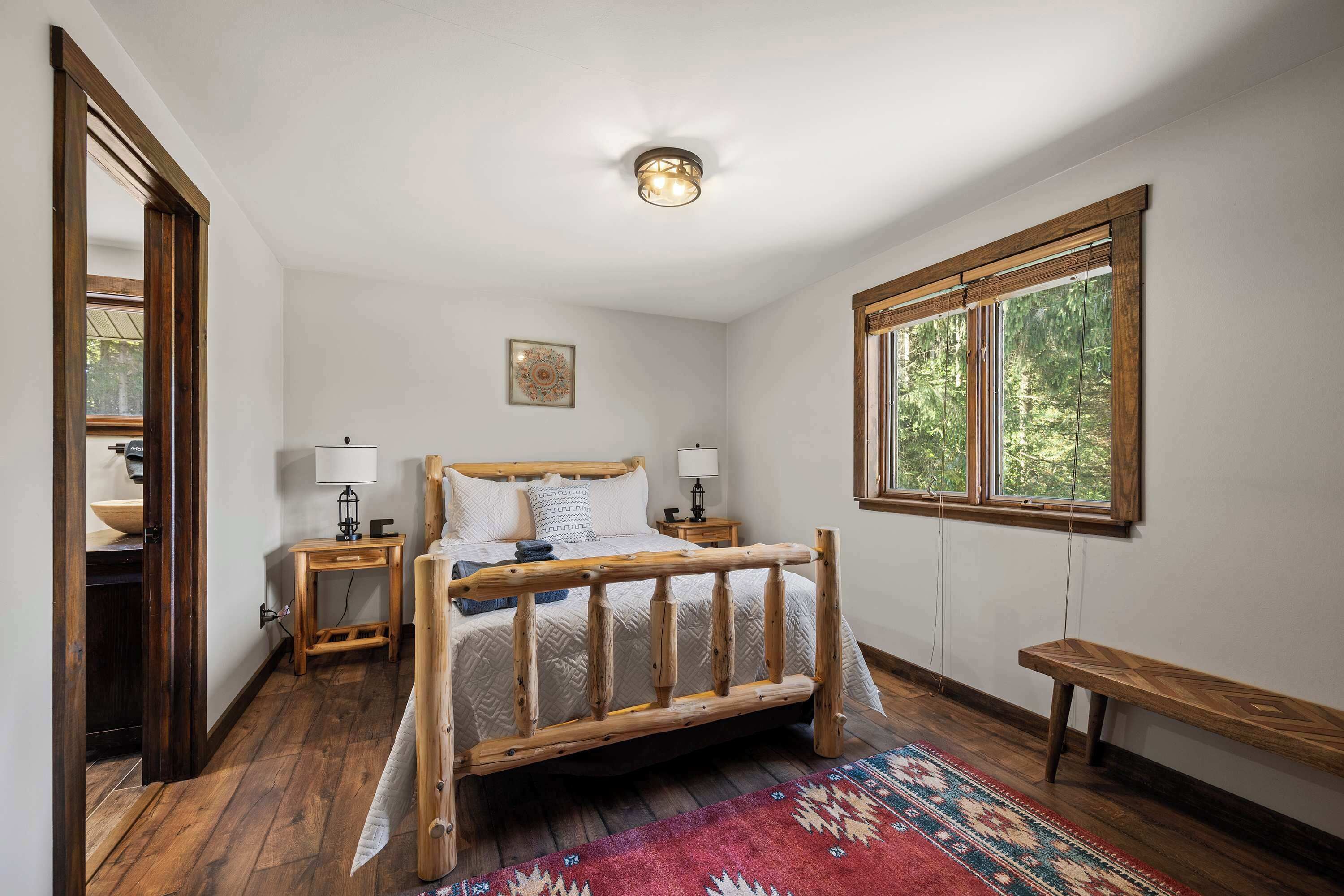 Une chambre à coucher avec un lit en bois, deux tables de chevet, un banc, une fenêtre donnant sur des arbres et un luminaire au plafond.