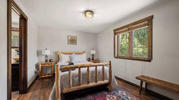 Une chambre à coucher avec un lit en bois, deux tables de chevet, un banc, une fenêtre donnant sur des arbres et un luminaire au plafond.