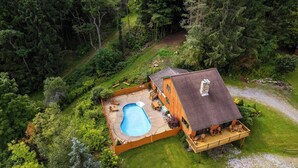 Una cabaña de madera con piscina, terraza y patio cercado, rodeada de un bosque denso.