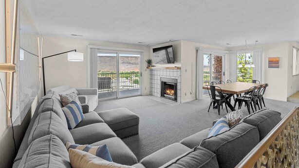 A spacious living room with a gray sectional sofa, a fireplace, a dining area with a wooden table and chairs, and large windows offering a view of the outdoors. - Cozy Condo - Desert Mesa View! (St. George)