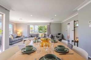 A dining area with a long table set for a meal, a sofa set in the background, and a view of the outdoors.