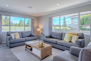 A modern living room with two grey sofas, a wooden coffee table, and large windows offering a view of a park and buildings.