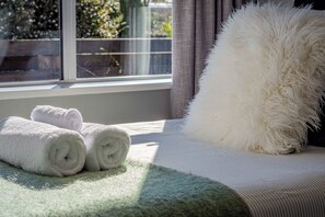 A neatly made bed with rolled white towels and a fluffy white pillow, positioned by a window with a view of greenery outside.