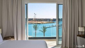 Quarto de hotel com cama, mesa de cabeceira, abajur e uma grande janela com vista para um porto e água.