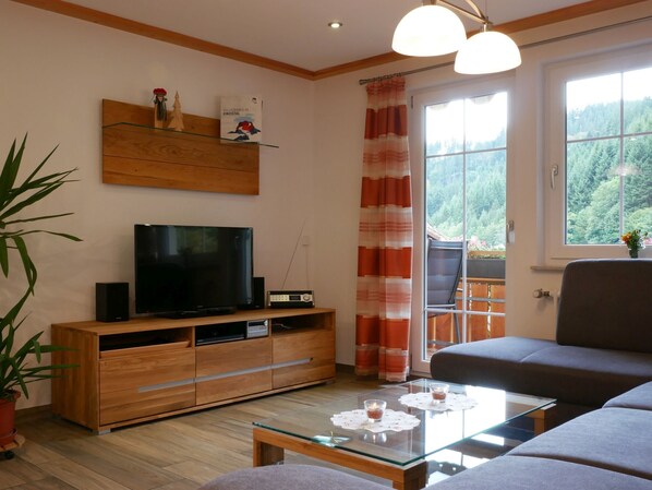 A living room with a TV, wooden entertainment unit, glass coffee table, sofa, and a view of a balcony with a railing. - Apartment 'Eulennest' with Shared Pool, Private Terrace and Wi-Fi (Wolfach)