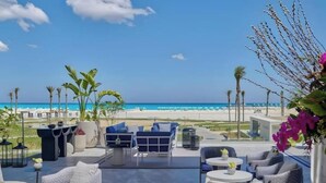 A beachside patio with outdoor seating, palm trees, and a view of the ocean. - Deluxe 1-BR Sea view Address Beach Marassi (El Alamein)