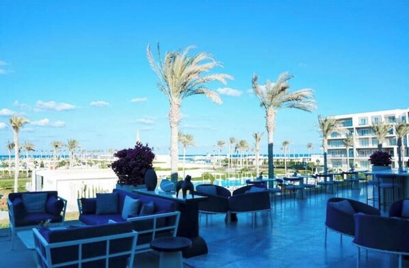 A poolside area with lounge chairs and tables, palm trees, and a view of a hotel building.