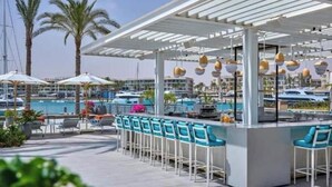 A covered outdoor bar area with blue bar stools, a pool with boats, and palm trees in the background.