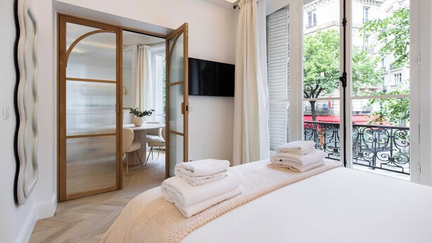 A hotel room with a bed, a TV, a balcony with a view of the city, and a door leading to a dining area. - Charming Flat - 2bd/6p - Passy / Eiffel Tower (Paris)