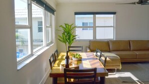 Un área de comedor con una mesa de madera, sillas, una planta y un sofá en una habitación luminosa con ventanas grandes.