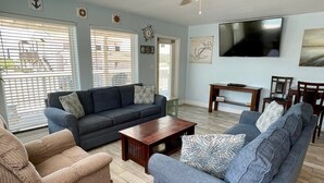 A living room with a dark blue sofa, a beige armchair, and a blue armchair. There is a wooden coffee table, a TV mounted on the wall, and a ceiling fan. - Beachgate Condo Suites And Hotel 211+212+213 3 Bedroom Condo (Port Aransas)