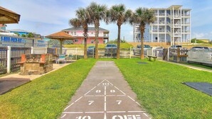 A shuffleboard court with a clear path leading to a building with balconies and a fenced area with palm trees. - Beachgate Condo Suites And Hotel 211+212+213 3 Bedroom Condo (Port Aransas)