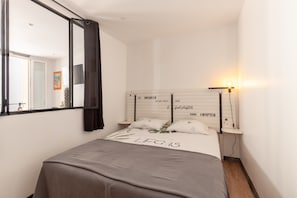 A bedroom with a bed, a bedside table, a nightstand, a mirror, and a wall-mounted light.