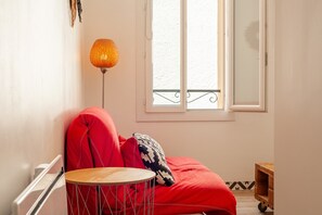 Une pièce avec un canapé rouge, une petite table en bois, un lampadaire et une fenêtre donnant sur un balcon.