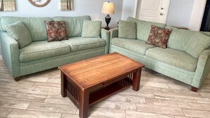 A living room with two green sofas, a wooden coffee table, and decorative wall art. - Beachgate Condo Suites And Hotel 516+517a+517b 3 Bedroom Condo (Port Aransas)