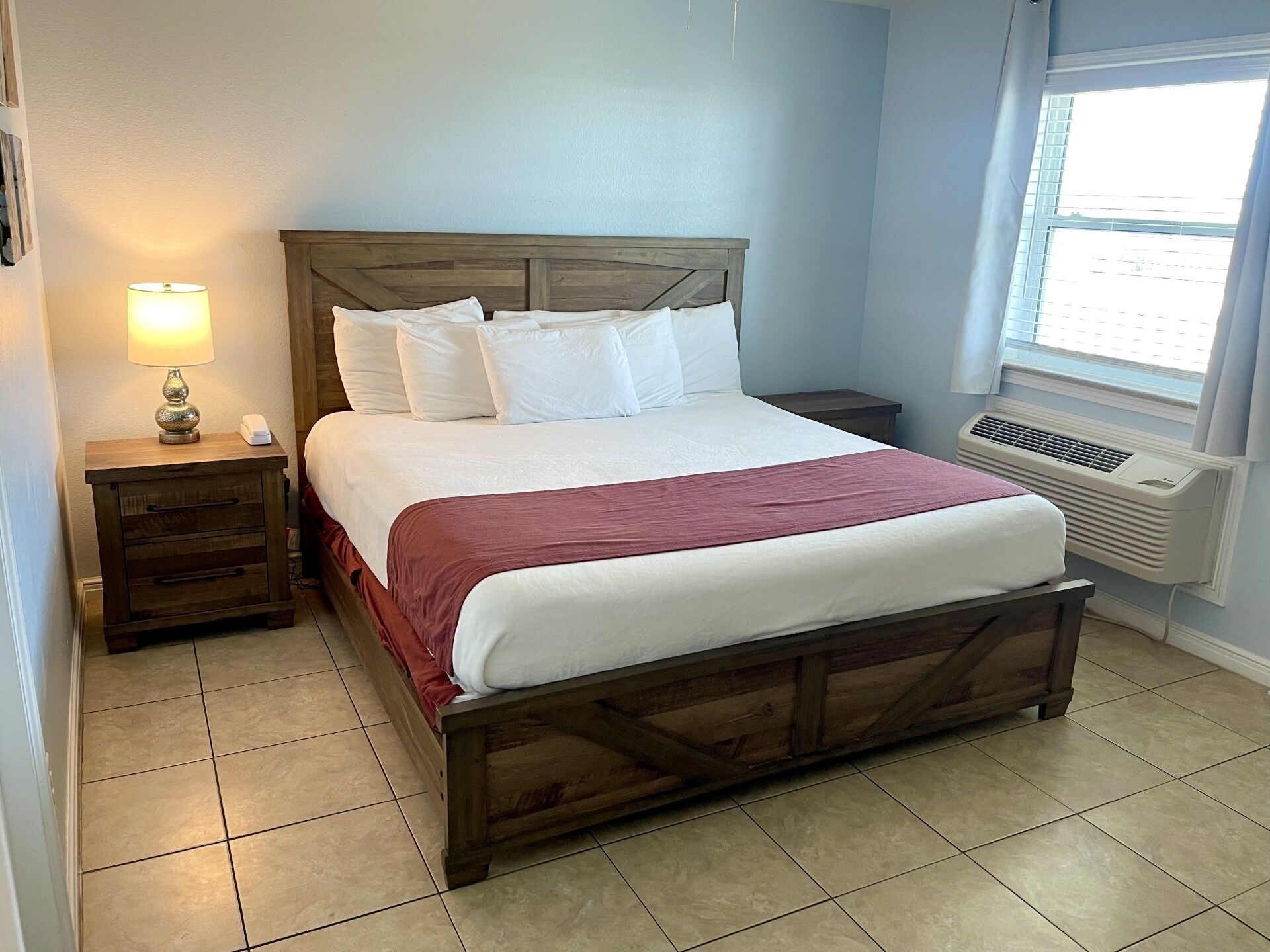 Una cama bien hecha con tablero de madera, una lámpara de noche, una mesita de noche y una ventana con aire acondicionado.