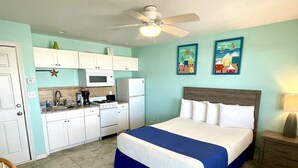 A compact hotel room with a kitchenette, a bed with a blue bedspread, and two wall art pieces depicting tropical scenes. - Beachgate Condo Suites And Hotel 234+235+236 3 Bedroom Condo (Port Aransas)
