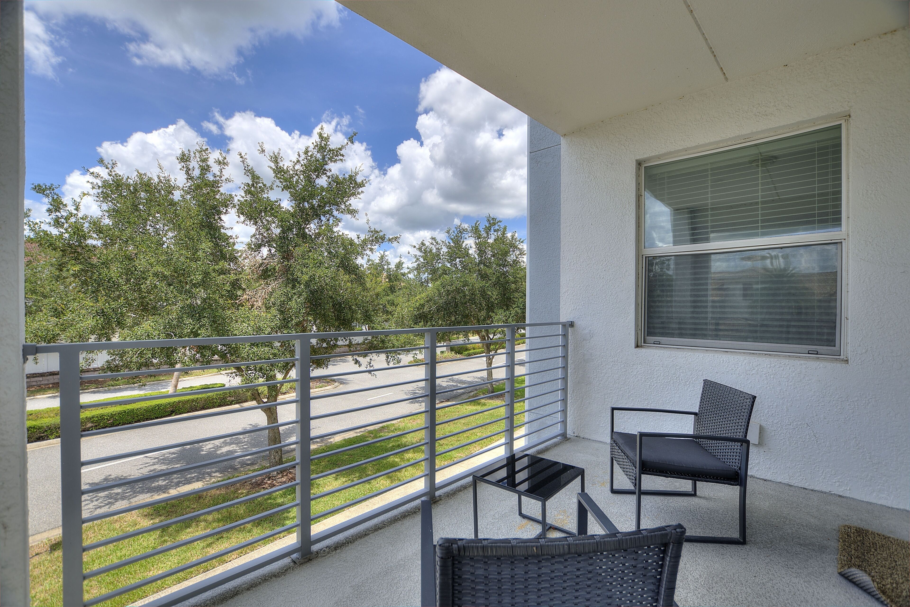 A balcony with a metal railing, two wicker chairs, a small table, and a window with blinds.