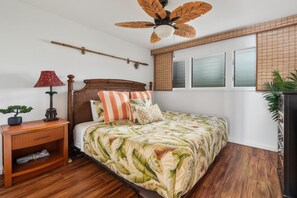 A bedroom with a bed, wooden nightstand, ceiling fan, and a window with blinds. - Big Island Kona Isle D3 1 Bedroom Condo (Kailua-Kona)