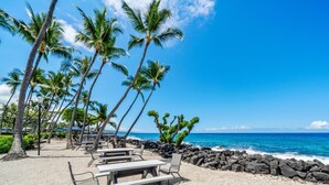A beachside dining area with tables and chairs, palm trees, and a view of the ocean. - Big Island Kona Isle D3 1 Bedroom Condo (Kailua-Kona)