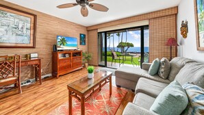 A living room with a wooden floor, a ceiling fan, a sofa, a coffee table, and a TV. - Big Island Kona Isle D3 1 Bedroom Condo (Kailua-Kona)