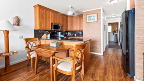 A modern kitchen with wooden cabinets, a microwave, and a refrigerator. There is a dining table with chairs and a ceiling fan above. - Big Island Kona Isle D3 1 Bedroom Condo (Kailua-Kona)