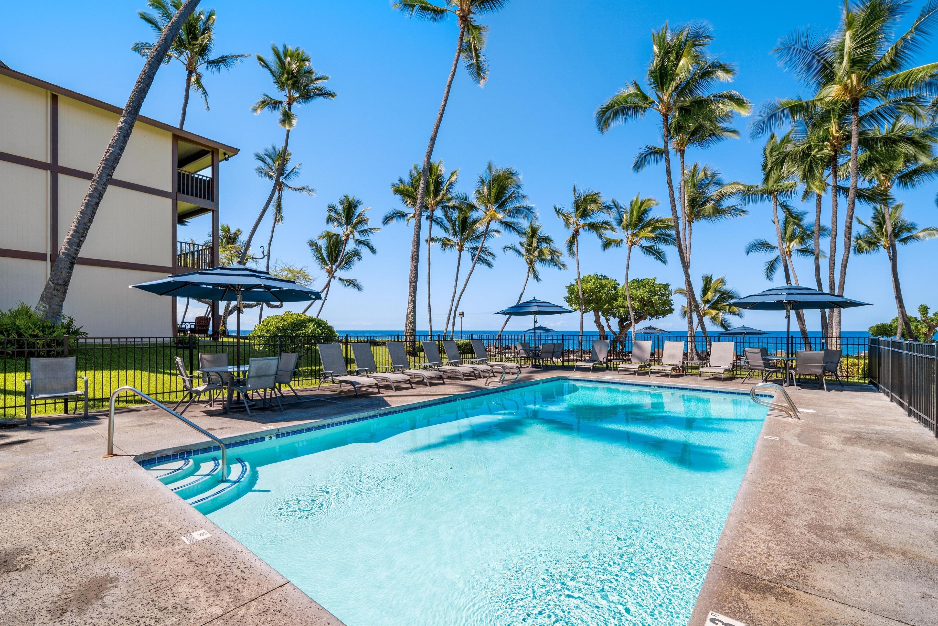 A pool area with lounge chairs and umbrellas, surrounded by palm trees and a building with a balcony.