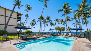 A pool area with lounge chairs and umbrellas, surrounded by palm trees and a building with a balcony. - Big Island Kona Isle D3 1 Bedroom Condo (Kailua-Kona)