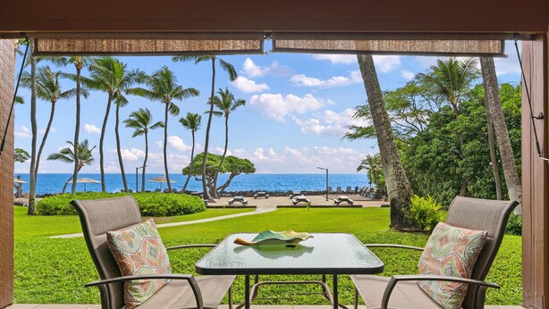 A patio with two chairs and a table, overlooking a beach with palm trees and a clear sky. - Big Island Kona Isle D3 1 Bedroom Condo (Kailua-Kona)