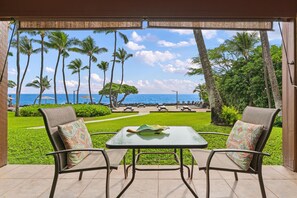A patio with two chairs and a table, overlooking a beach with palm trees and a clear sky. - Big Island Kona Isle D3 1 Bedroom Condo (Kailua-Kona)