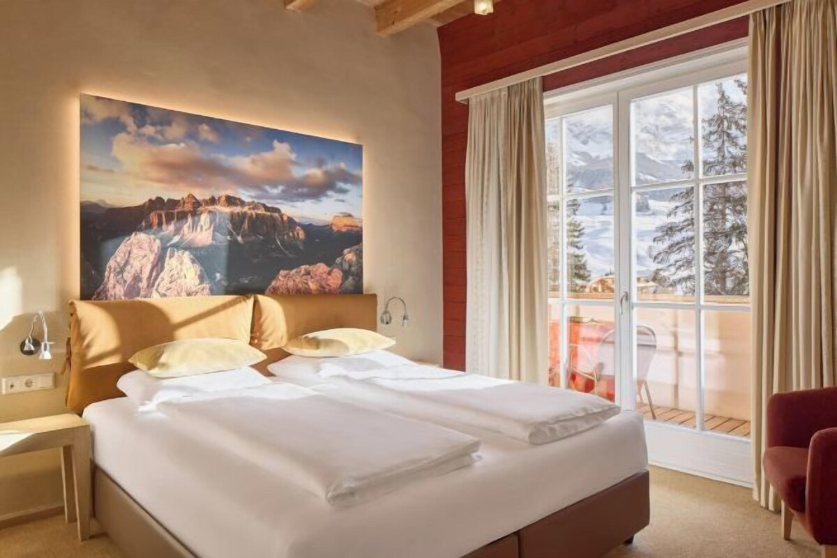 A hotel room with a large bed, a painting of a mountain landscape, a window with a view of trees, and a red chair.