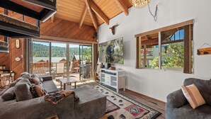 A spacious living area with a large sofa, a coffee table, and a rug with geometric patterns. There is a sliding glass door leading to an outdoor patio with a view of a forested area.